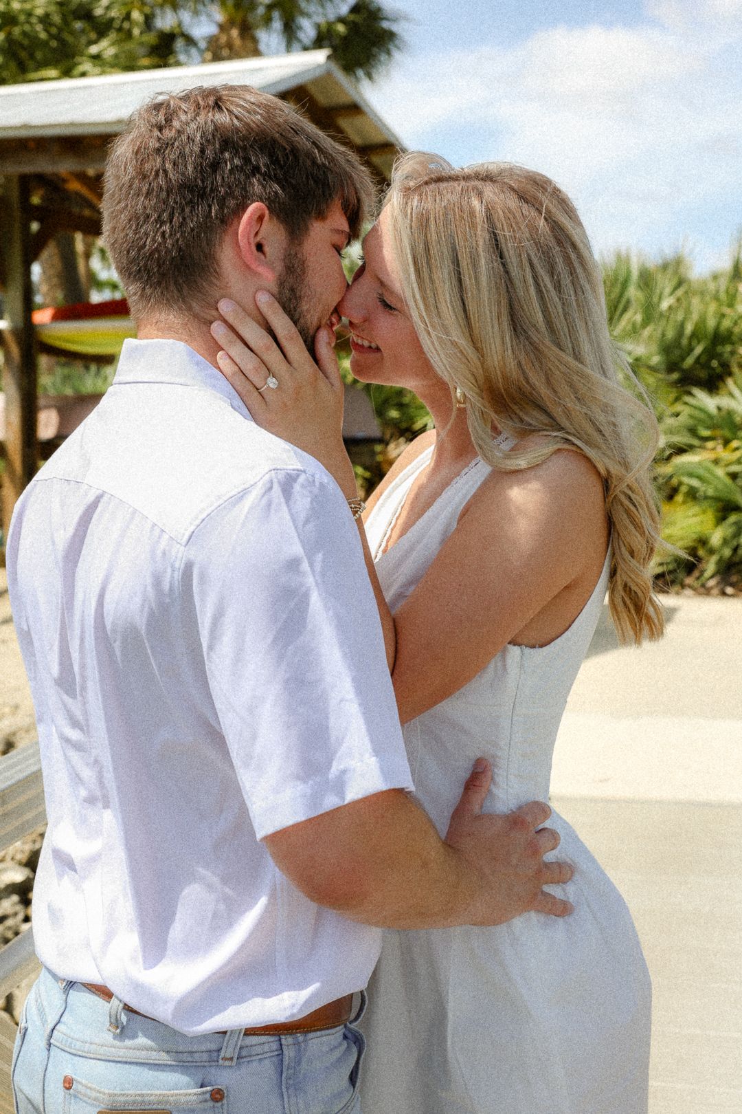 An engaged couple sharing a kiss outdoors, woman showing off her engagement ring.