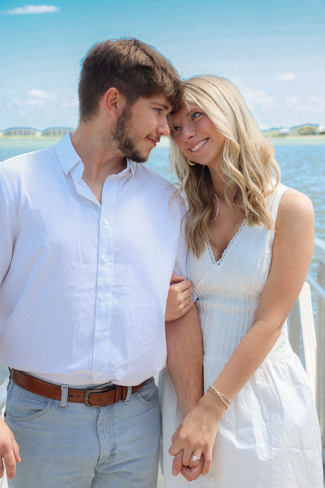 Engaged couple in white outfits touch foreheads tenderly while standing on dock with blue water behind them.
