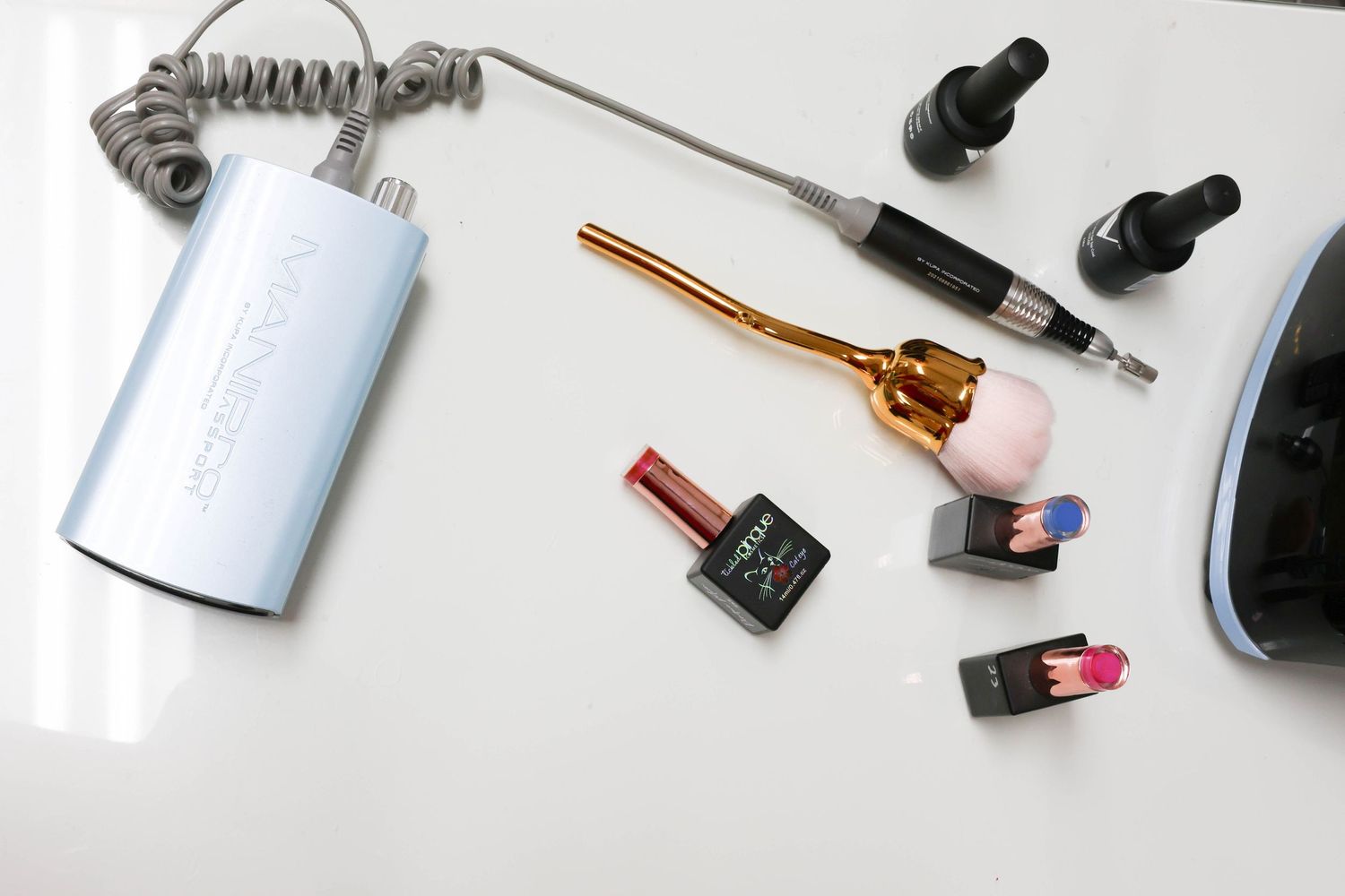 Nail salon tools including electric file, UV lamp, gel polishes, and brush on white surface.