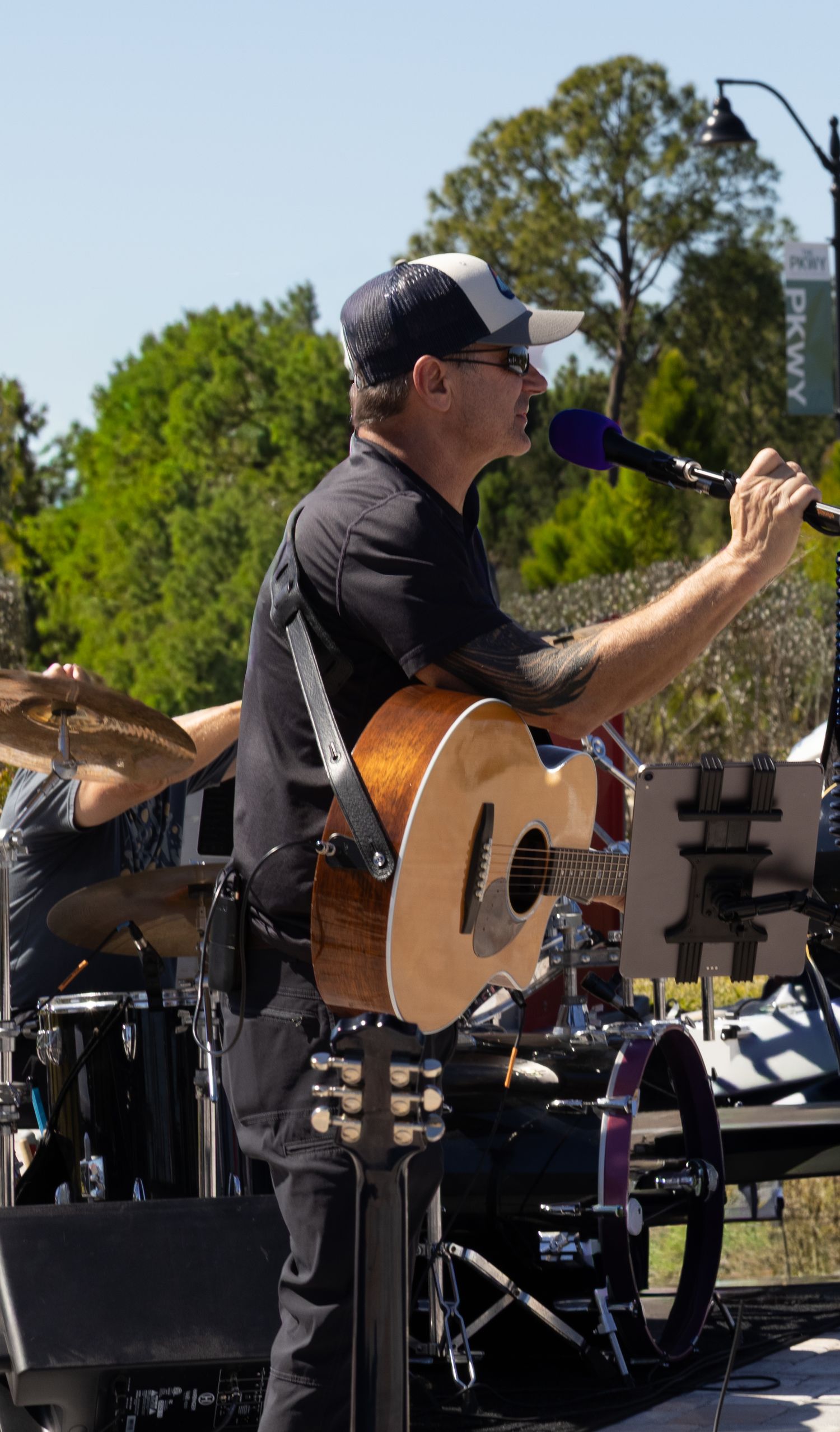 Male guitarist performing outdoors on stage, wearing black shirt and cap, singing into a microphone.