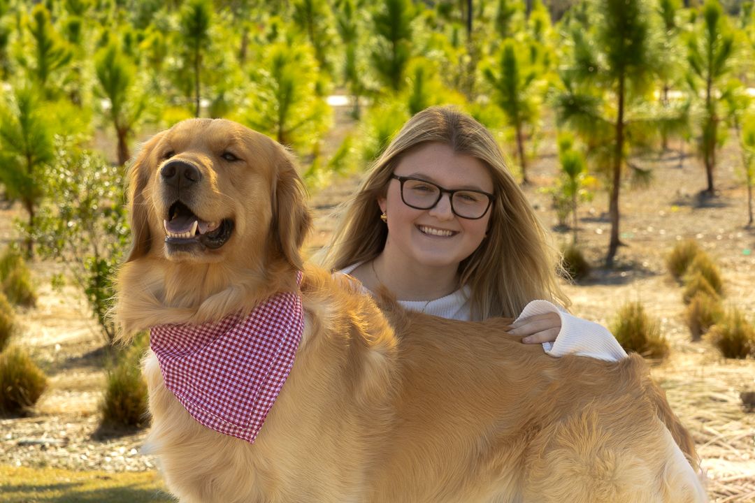 A smiling young woman with glasses embraces her golden retriever in a lush green tree nursery.