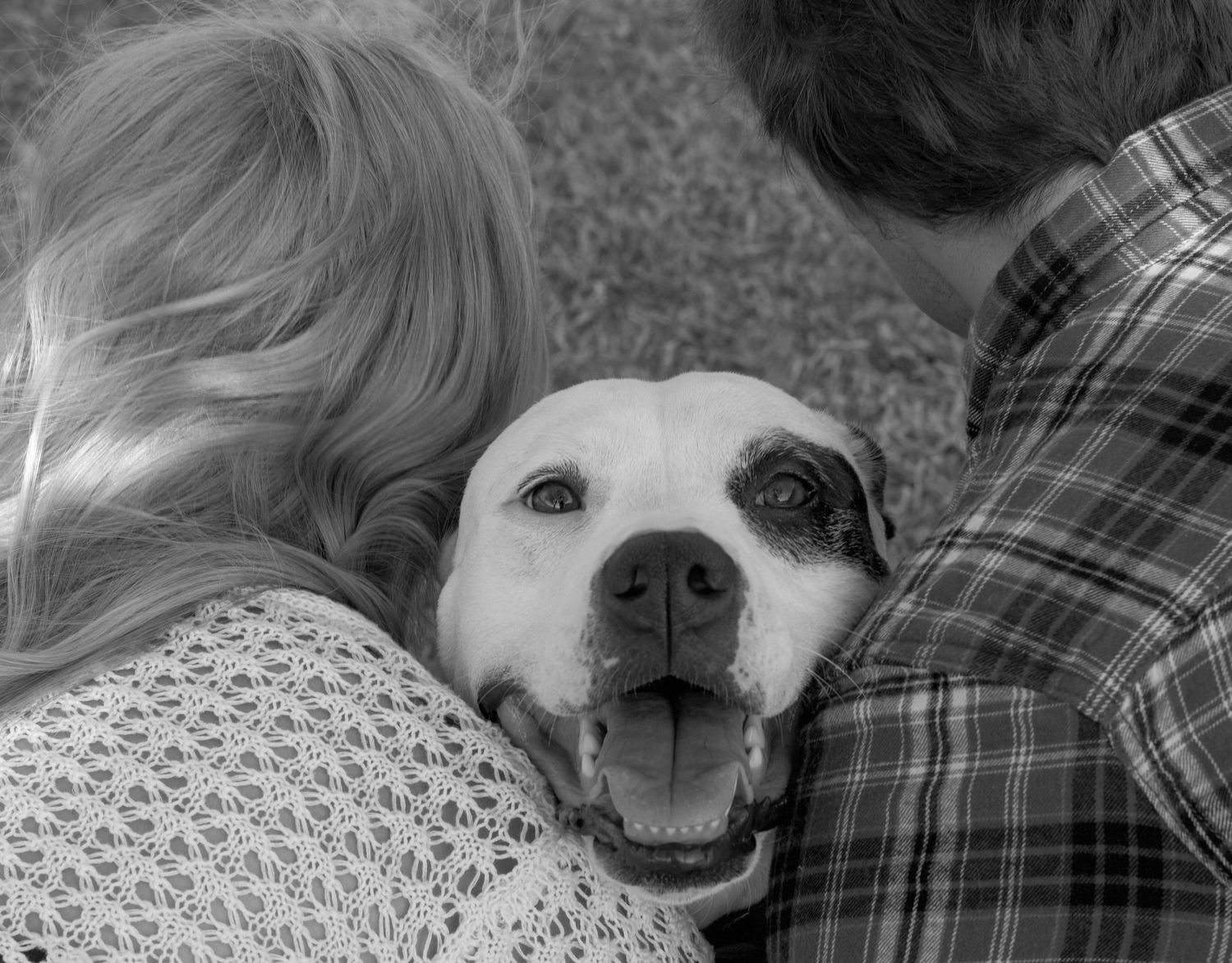 Black and white photo of a happy dog sandwiched between two people, tongue out and smiling at the camera.