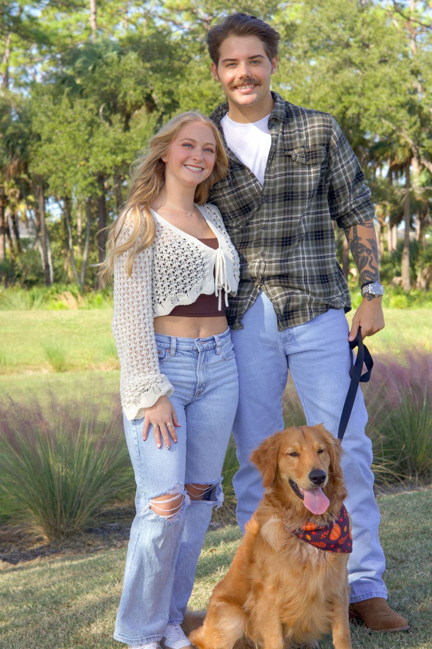 Smiling couple standing with a golden retriever wearing a bandana in a park.