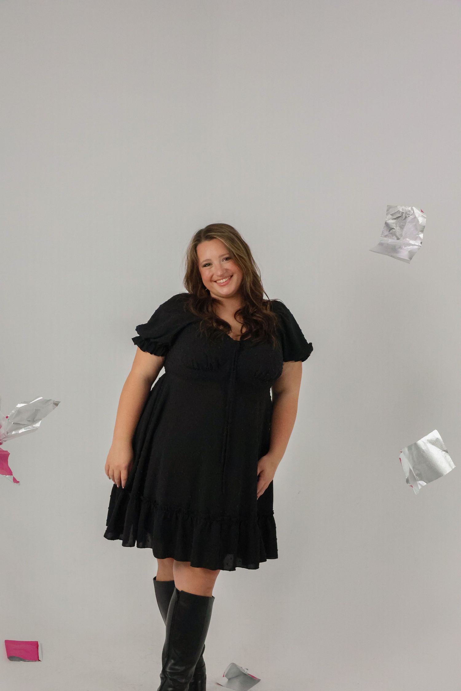 Woman in black dress and knee-high boots smiling as paper confetti falls around her.