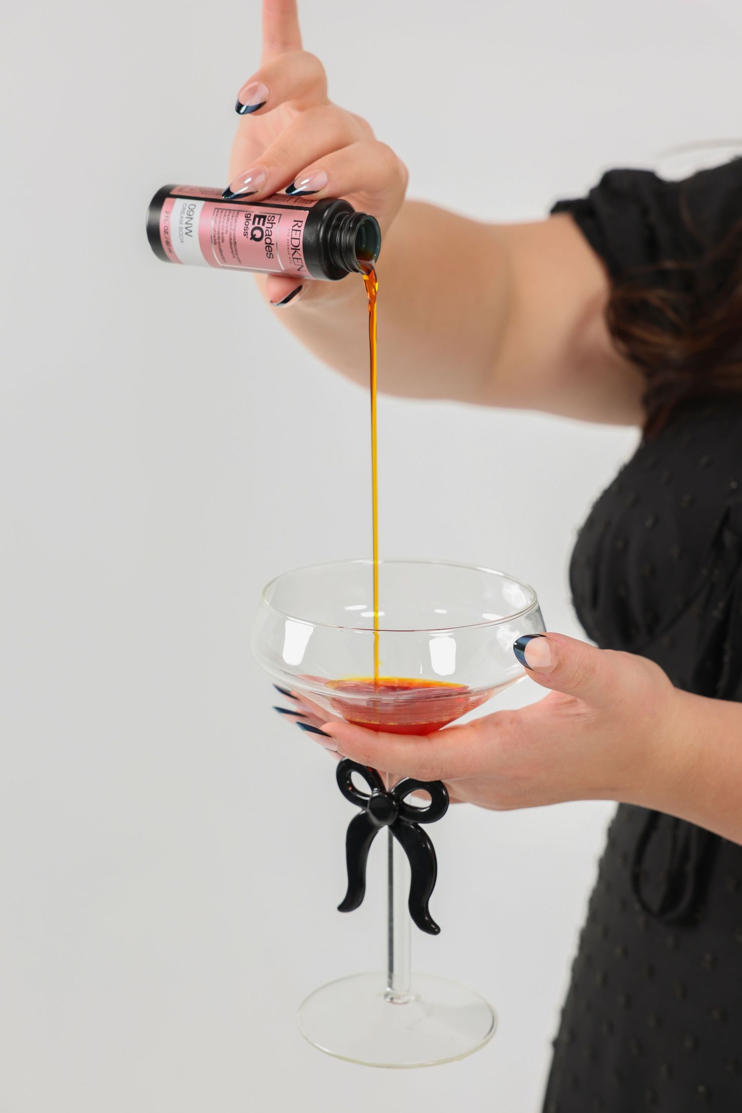 Hands with black nails pouring hair product into a decorative coupe glass with black bow.