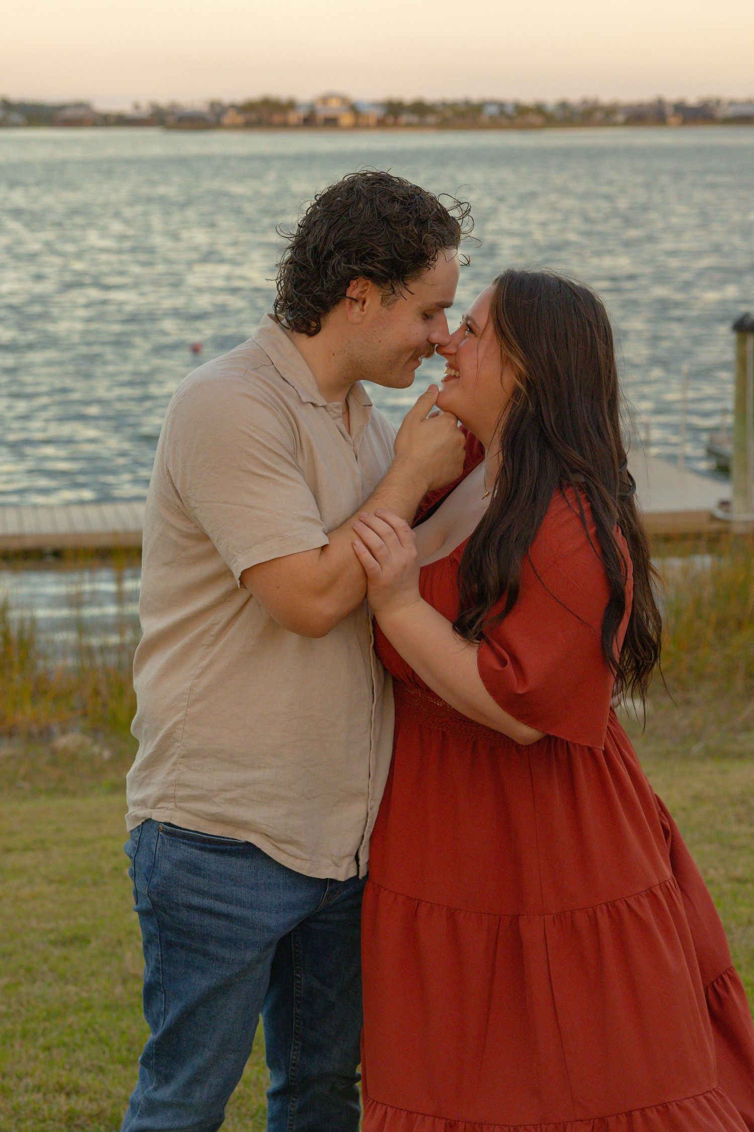 Couple sharing a romantic nose-to-nose moment by a lakeside at golden hour, dressed in red and beige.