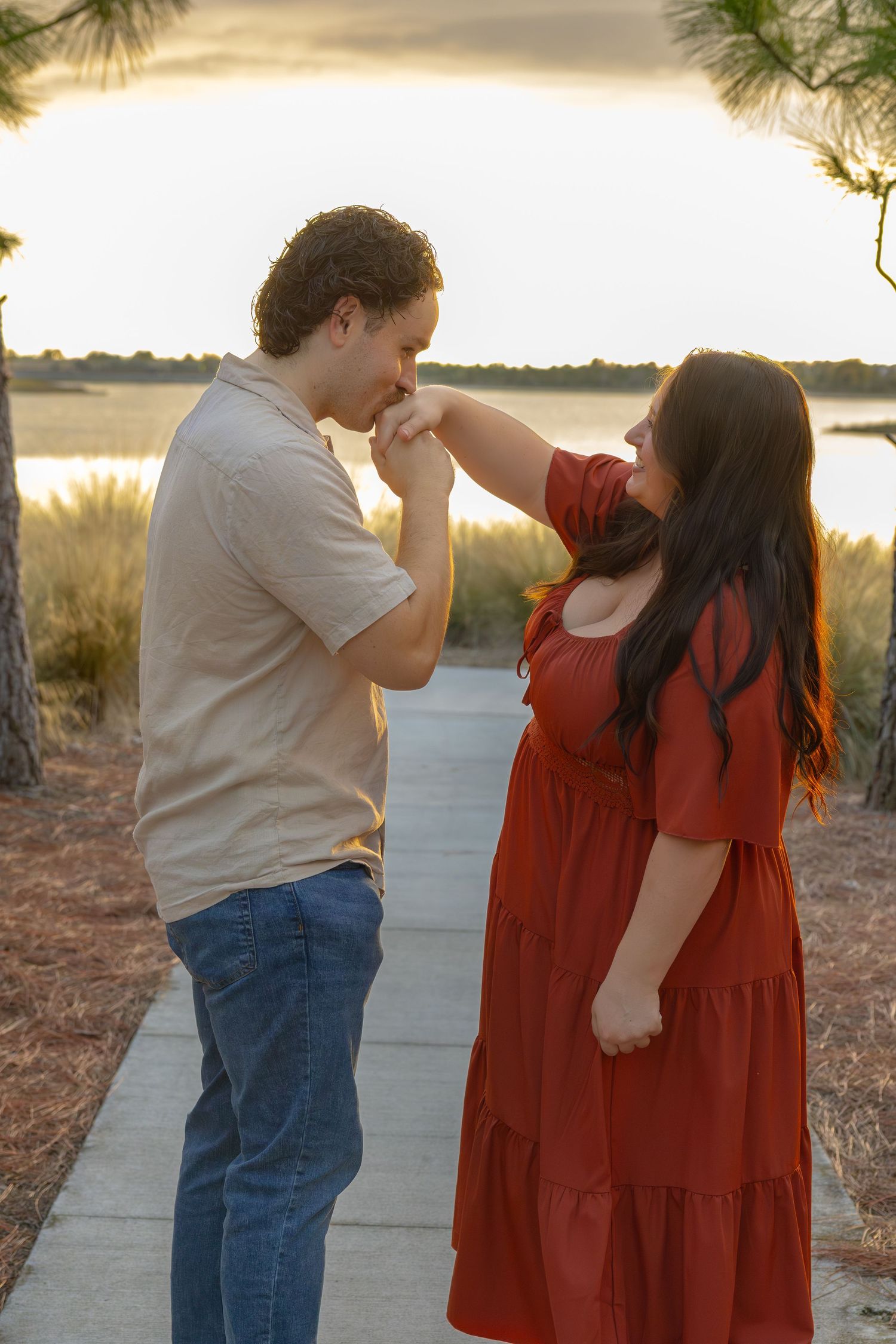 Man kisses woman's hand at sunset by a waterfront path, she wears a red dress.