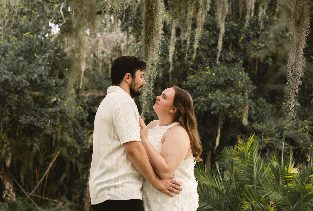 A happy couple embraces outdoors surrounded by lush greenery and Spanish moss-draped trees.