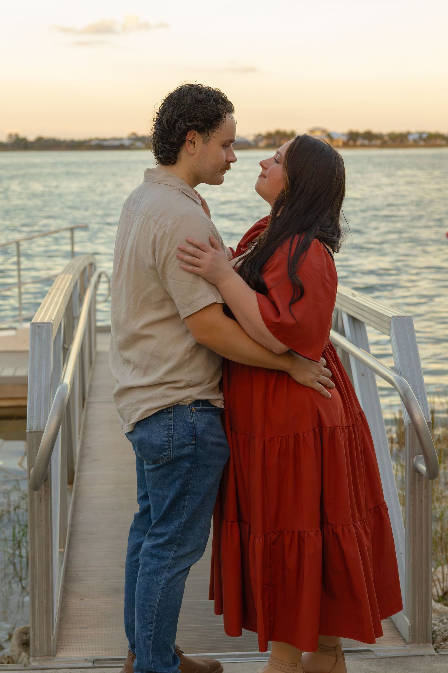 Romantic couple embraces on a waterfront dock at sunset, gazing into each other's eyes.