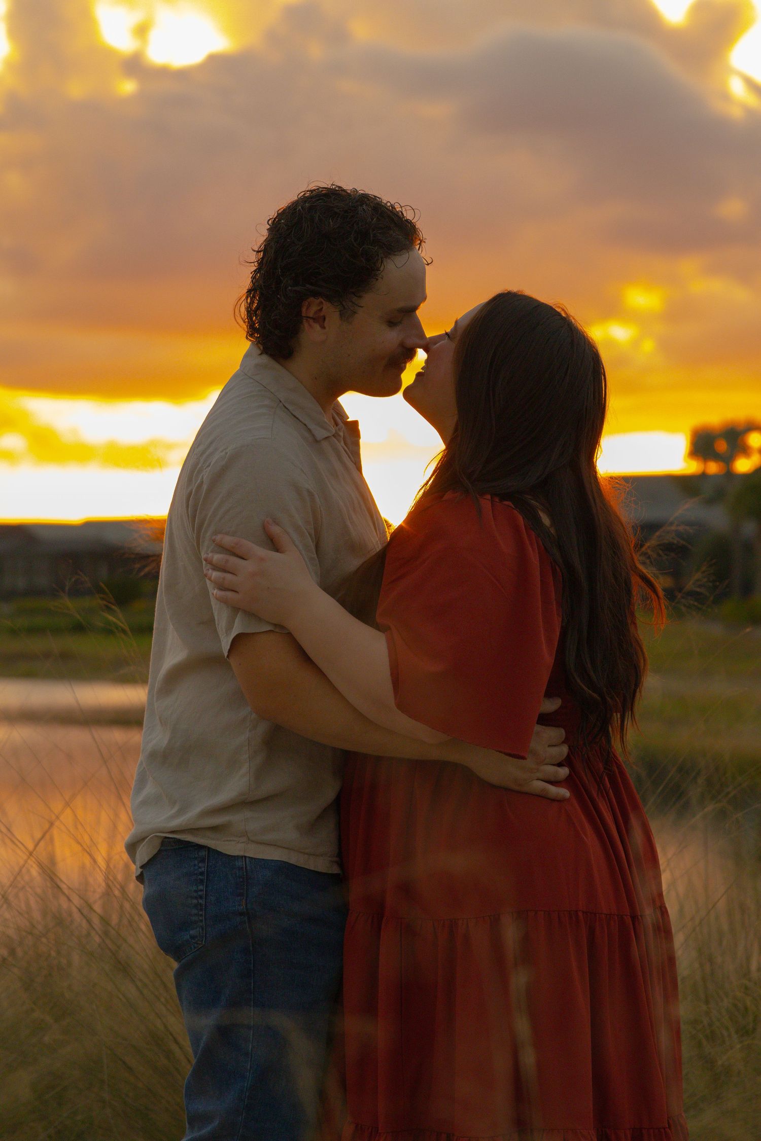 Couple sharing a romantic kiss silhouetted against a stunning golden sunset sky by the water.