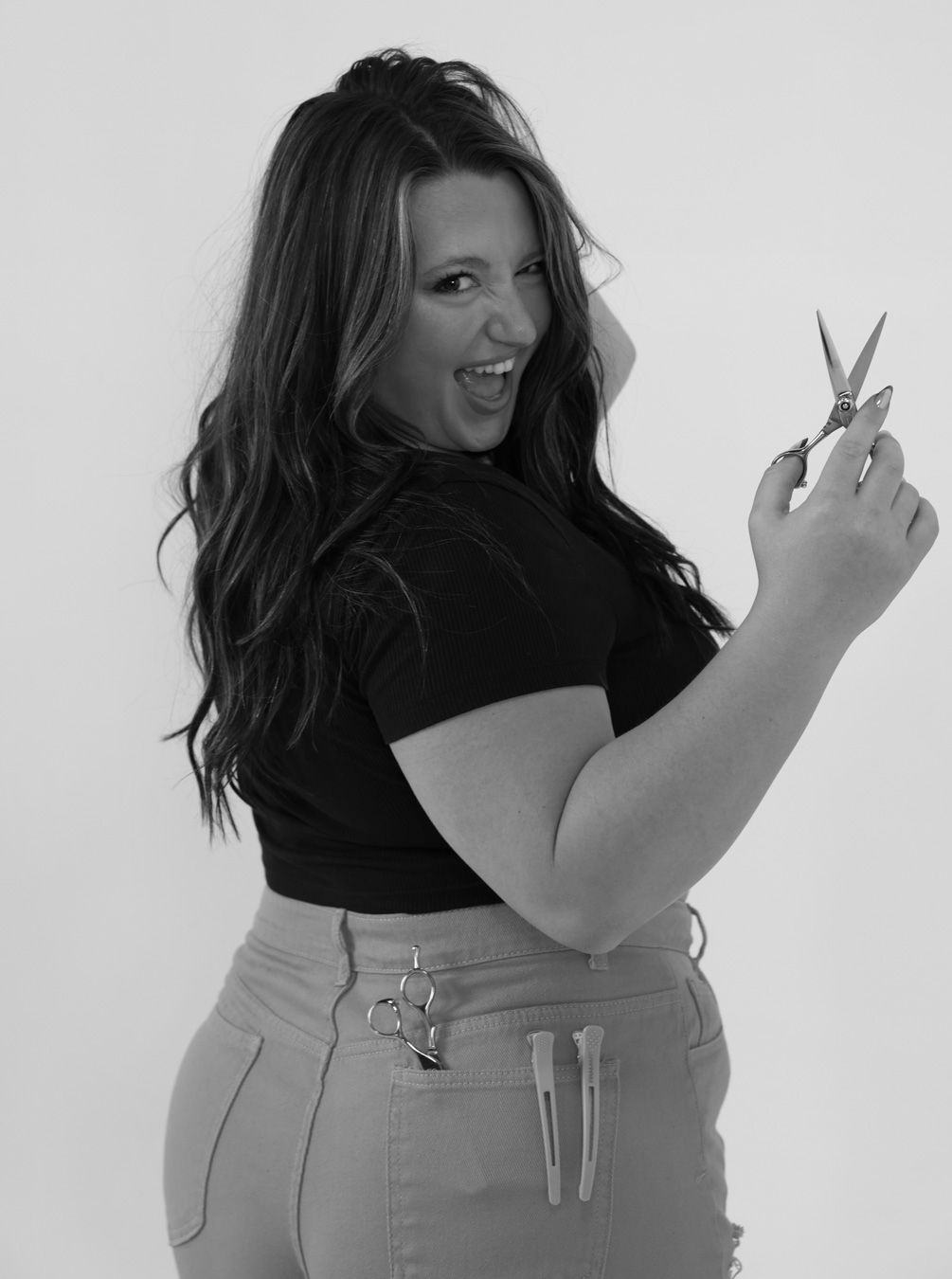 Woman in black top and jeans posing with scissors in black and white photo.