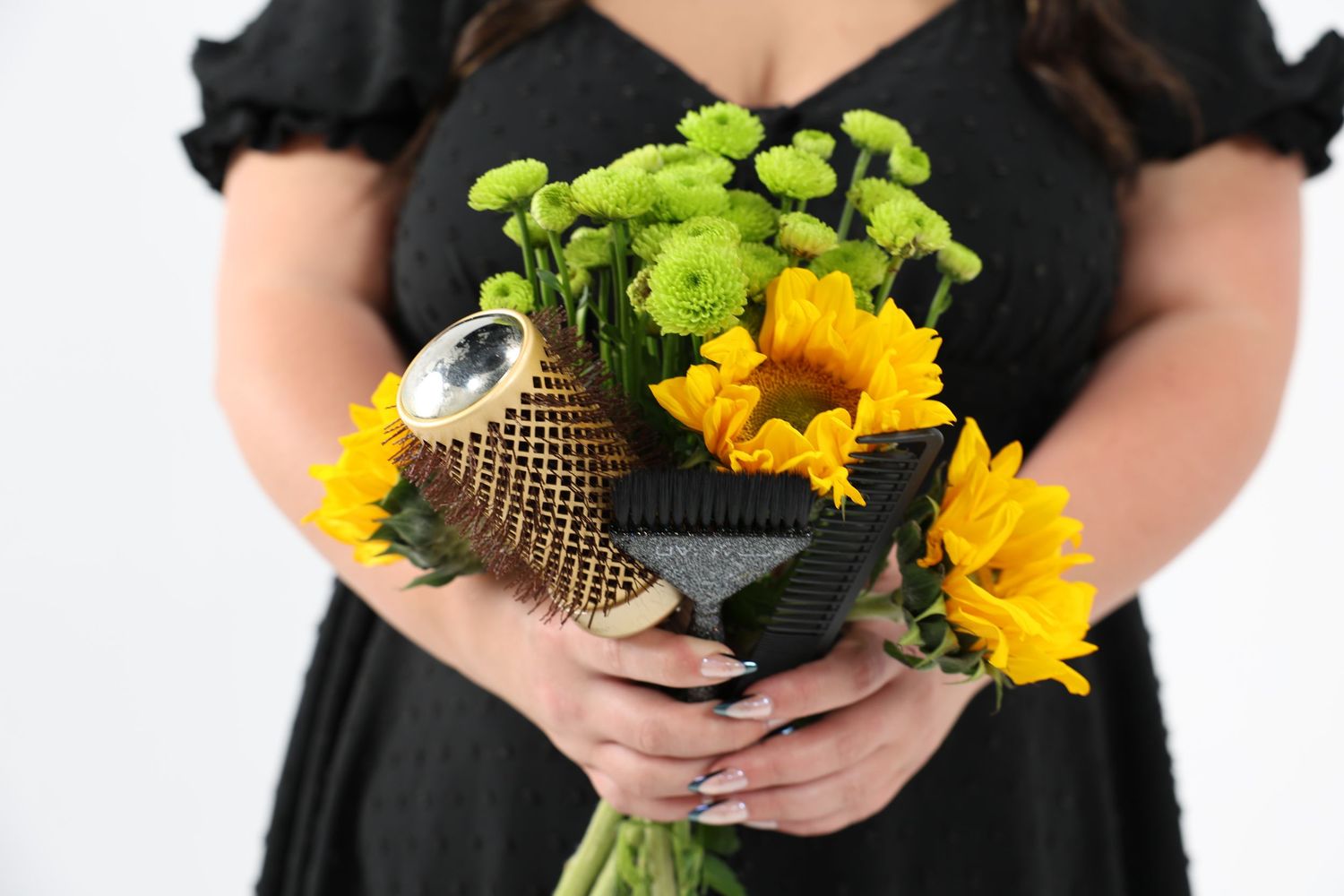 Close-up of hands holding a bouquet of sunflowers and green flowers with a round brush tucked in.
