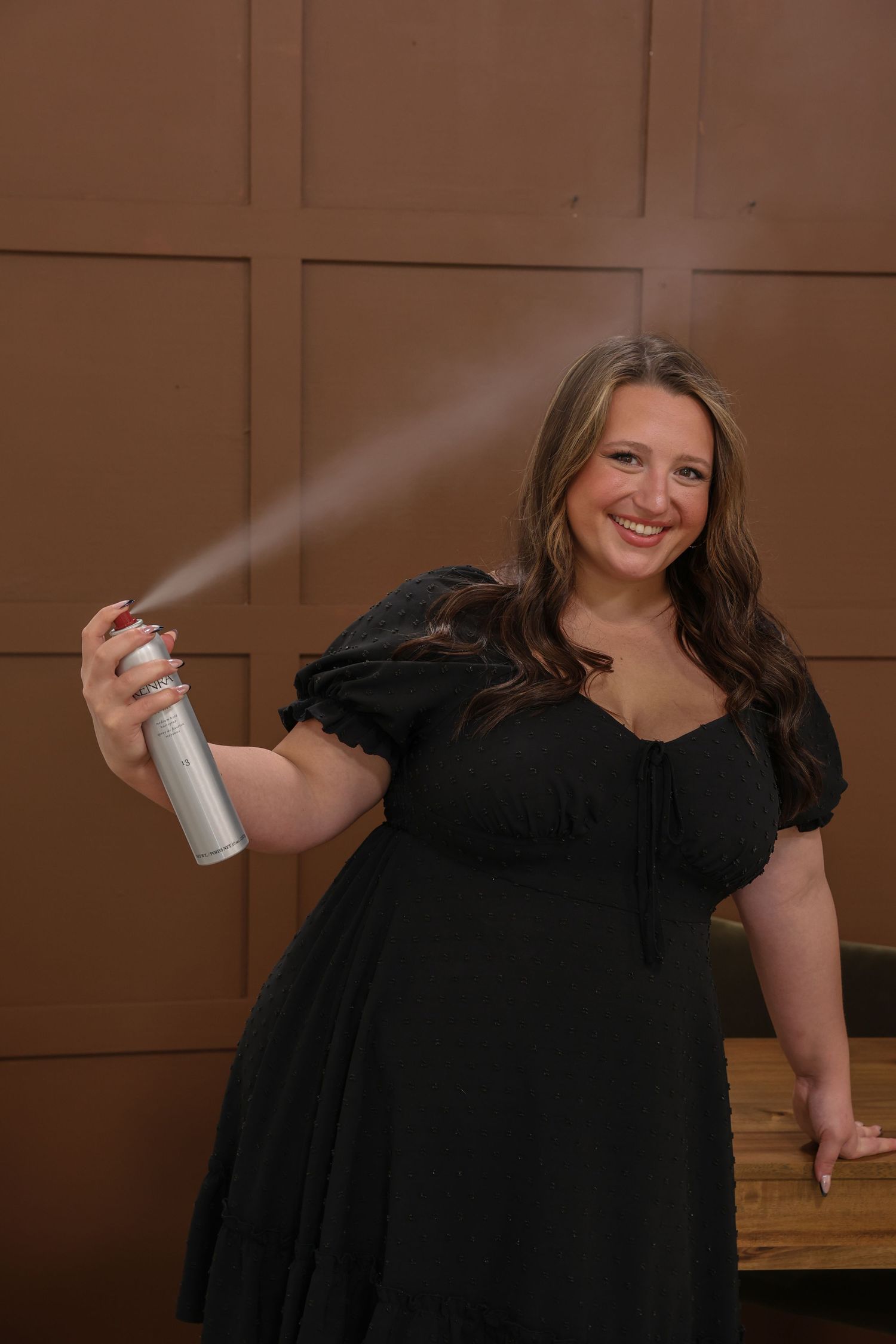 Woman in black dress smiling while spraying hairspray on brown paneled background.