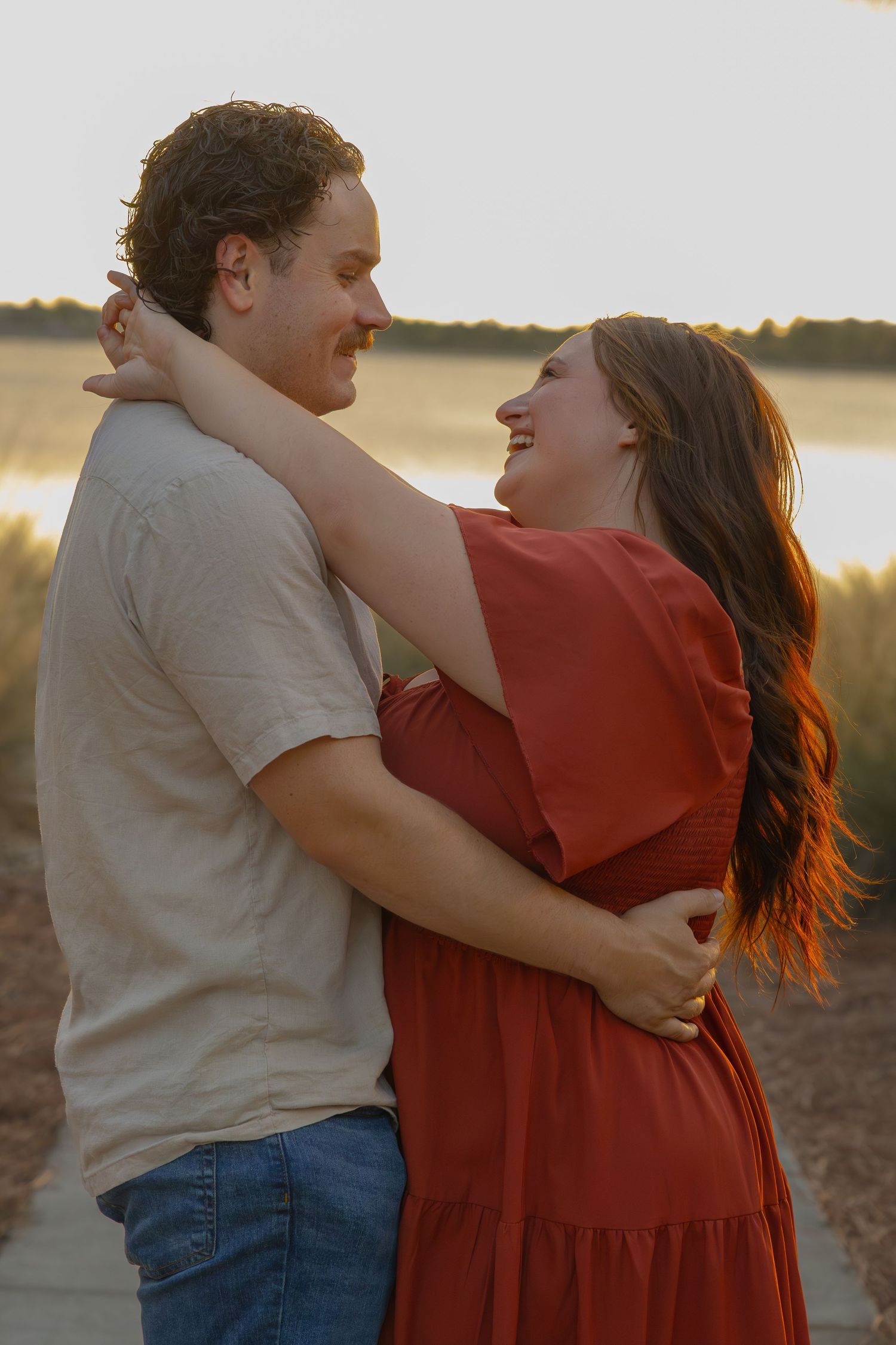 A happy couple embraces and laughs together at sunset near a waterfront, glowing in warm golden light.