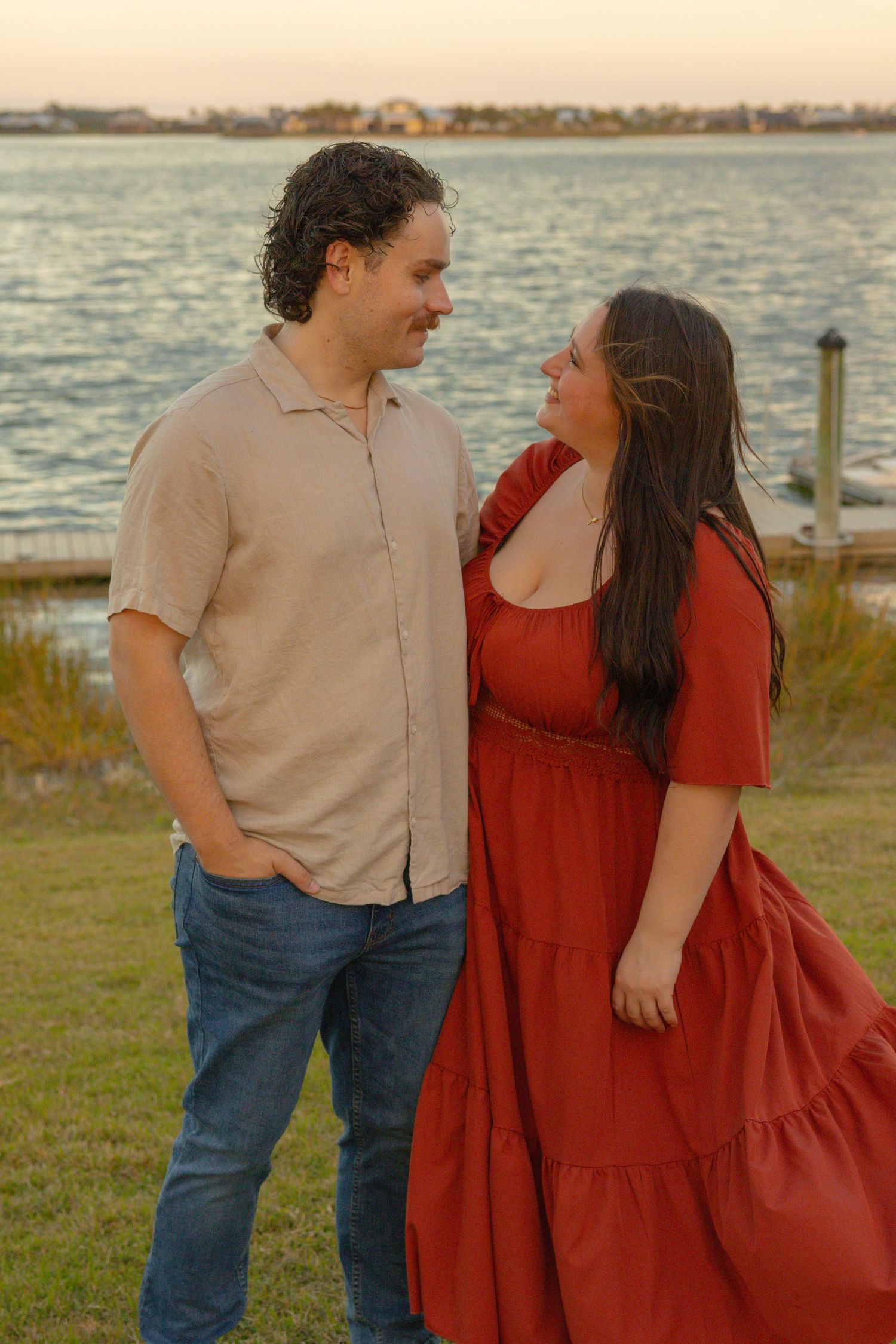 Romantic couple sharing a tender moment by the water at sunset, woman in red dress and man in beige shirt.