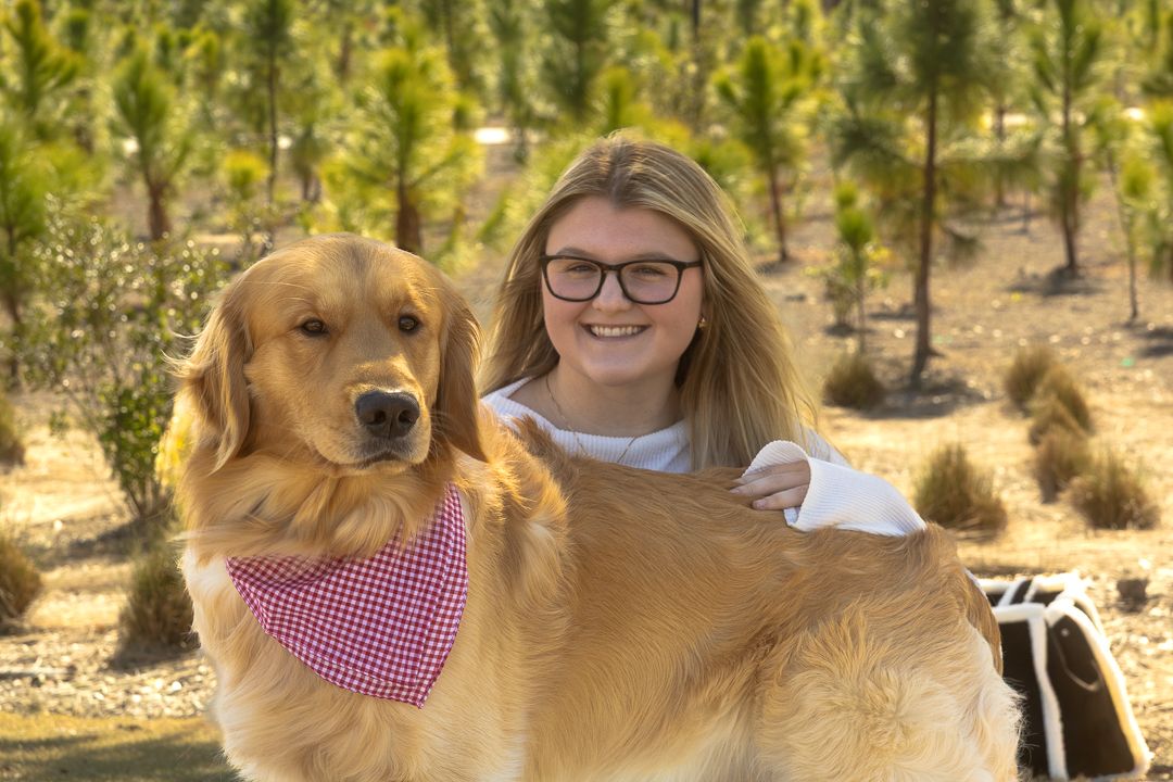 A cheerful young woman hugs her golden retriever with a pink bandana in a tree nursery setting.