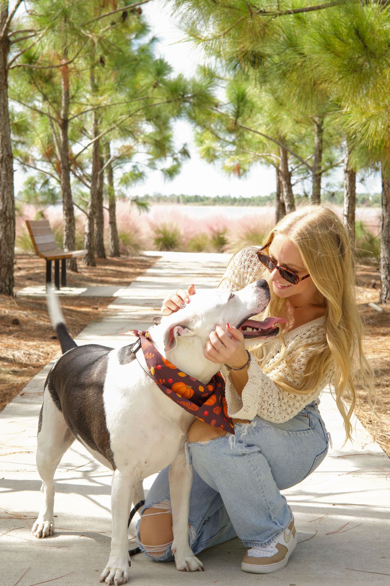 Blonde woman in sunglasses crouching on a park path, sharing a sweet moment with her dog wearing a bandana.