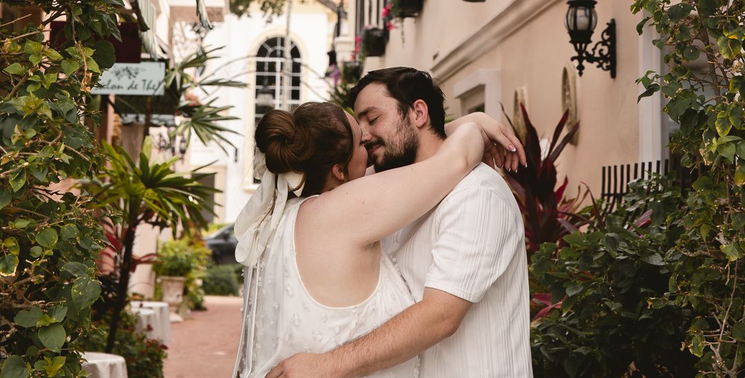 Couple sharing a romantic embrace and kiss outdoors among lush tropical greenery near a white building.