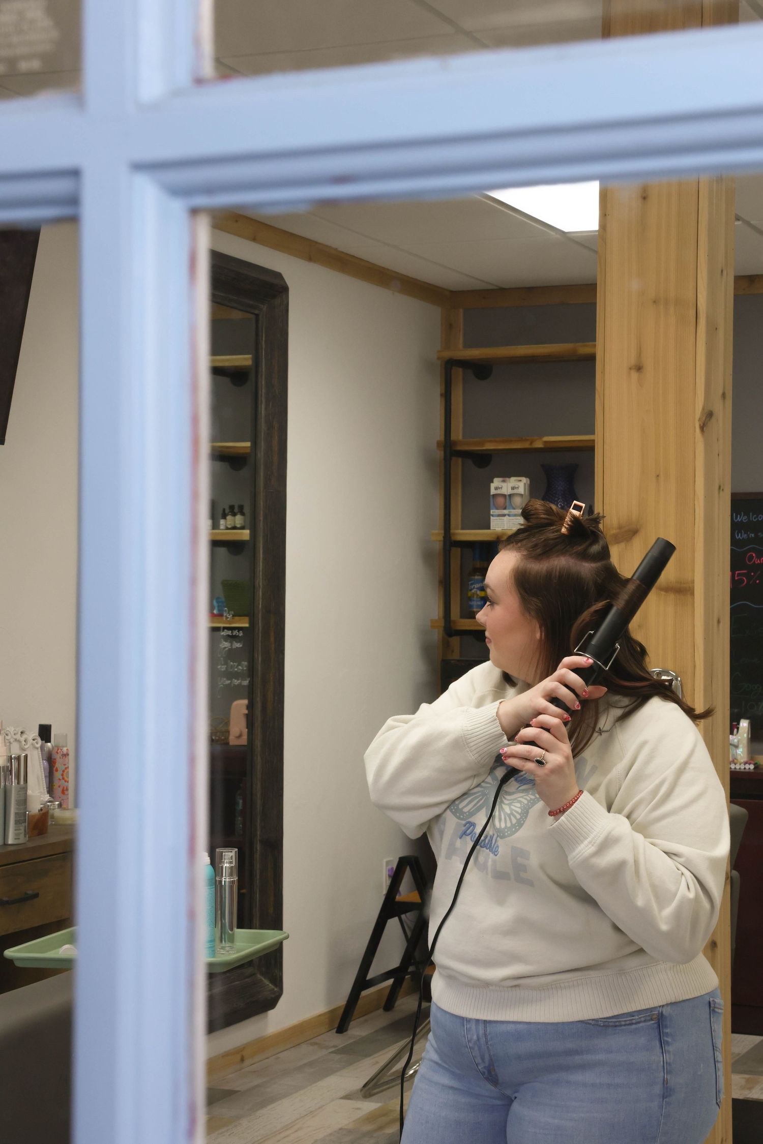 Woman using a curling iron on her hair viewed through a window in a modern hair salon.
