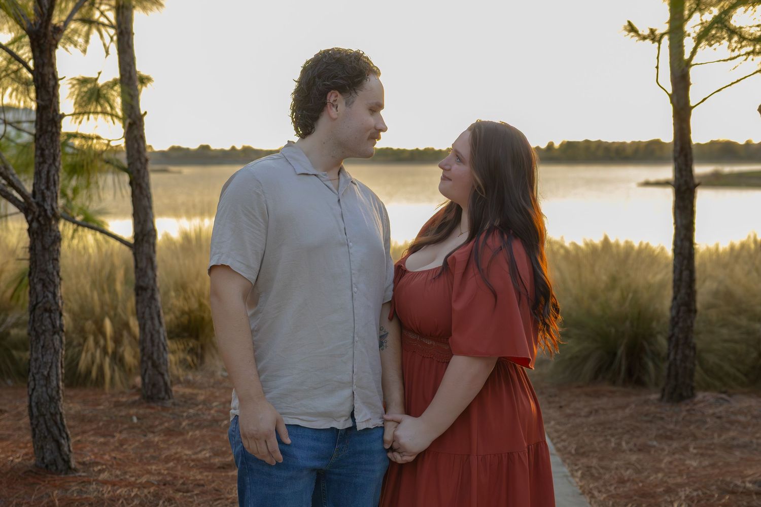 A smiling couple stands together at golden hour near a waterfront surrounded by pine trees.