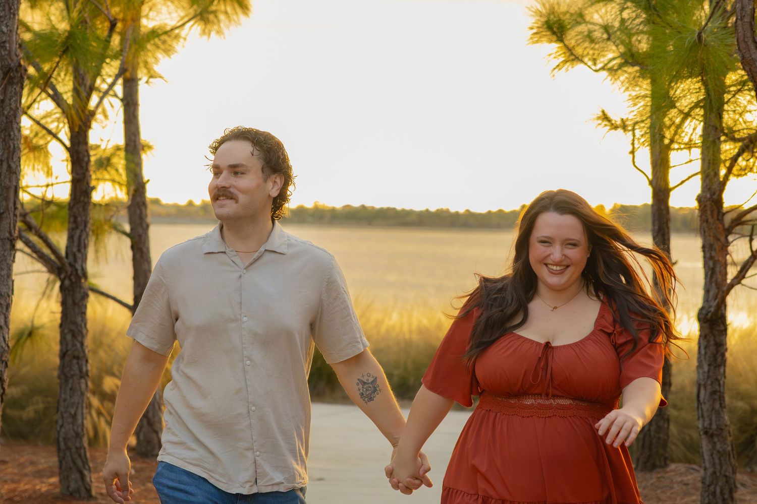 Couple holding hands walking outdoors near a lake at golden hour, woman in red dress, man in casual shirt.