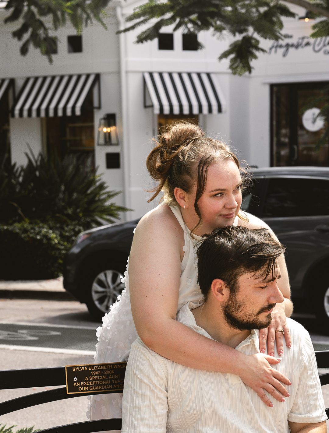 A young couple poses outdoors near a boutique storefront, woman resting on mans shoulders, both wearing white outfits.