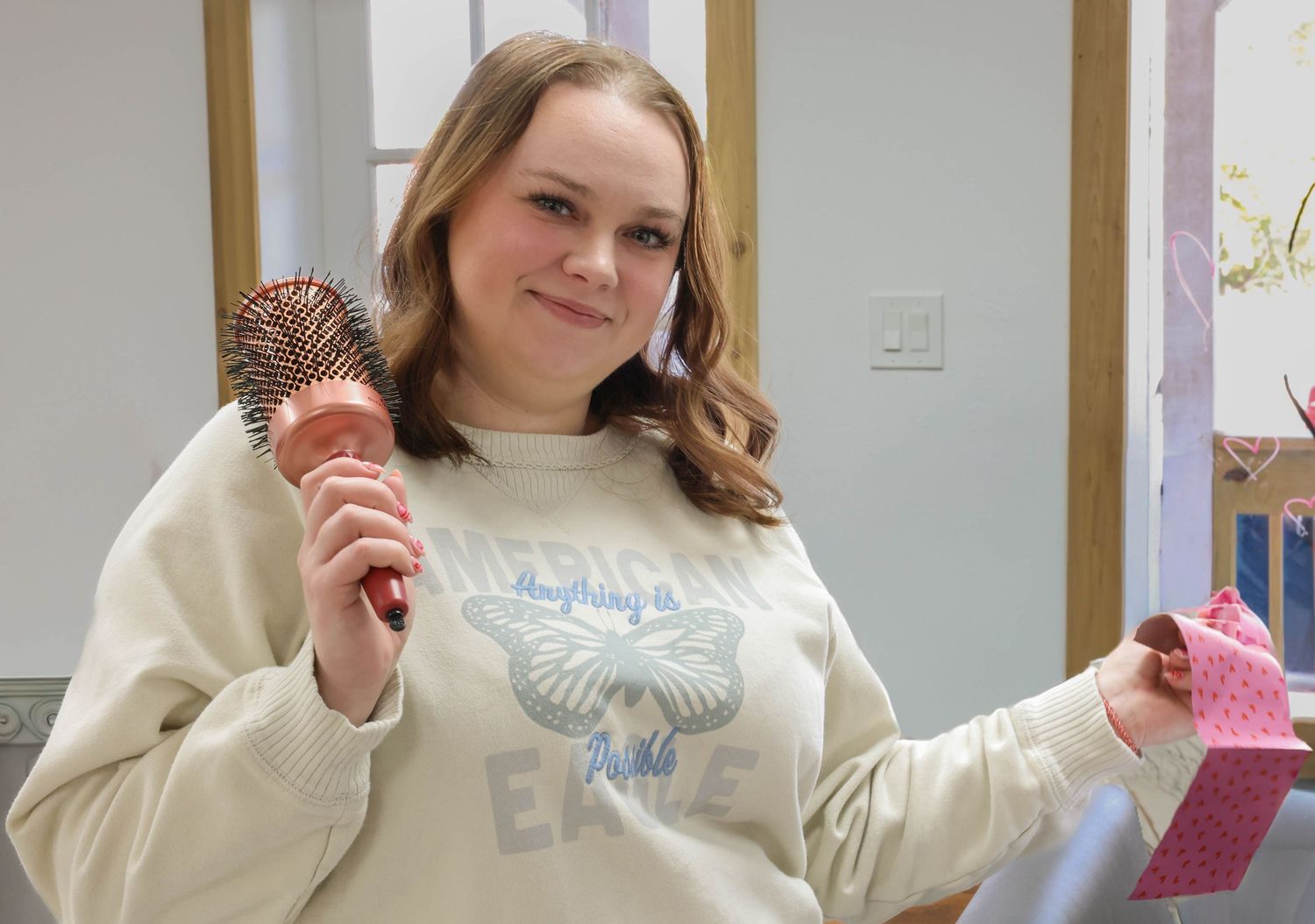 Young woman in cream sweatshirt smiling while holding a hair brush and pink gift box in a salon.