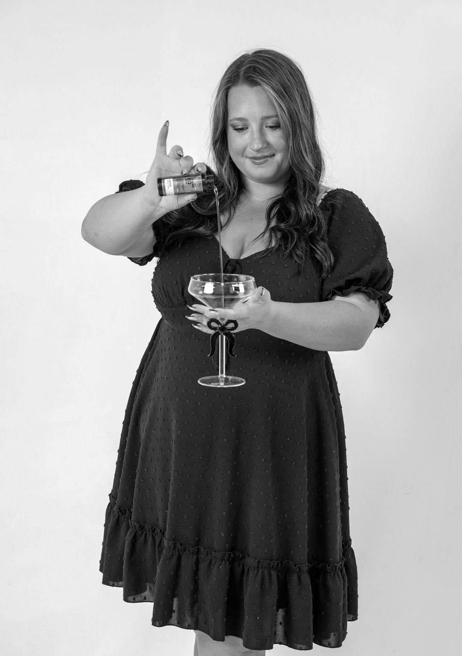 Black and white photo of woman in black dress pouring liquid into a bow-tied coupe glass.