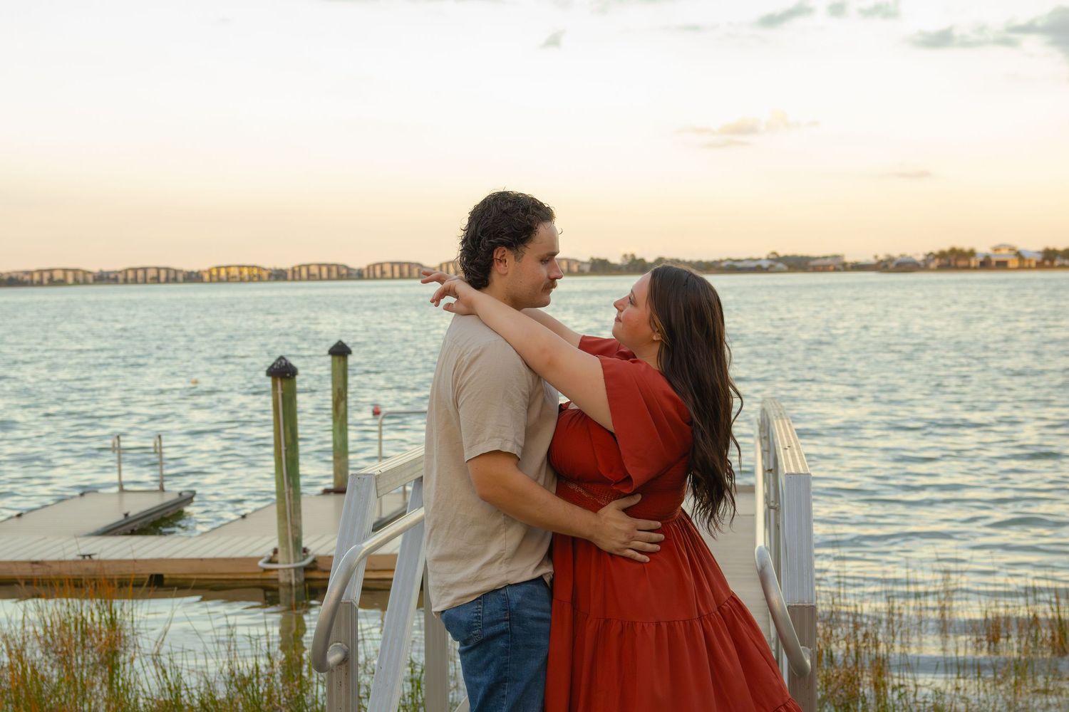 Couple embracing on a waterfront dock at sunset, woman in flowing red dress and man in beige shirt.