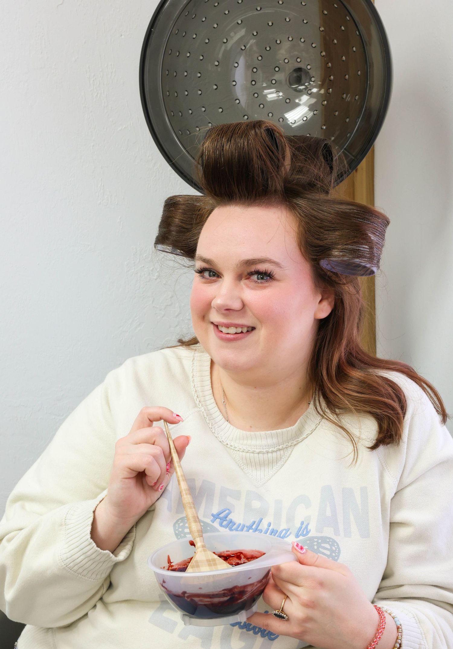 Woman in American Eagle sweatshirt eating from bowl with large hair rollers in her hair.
