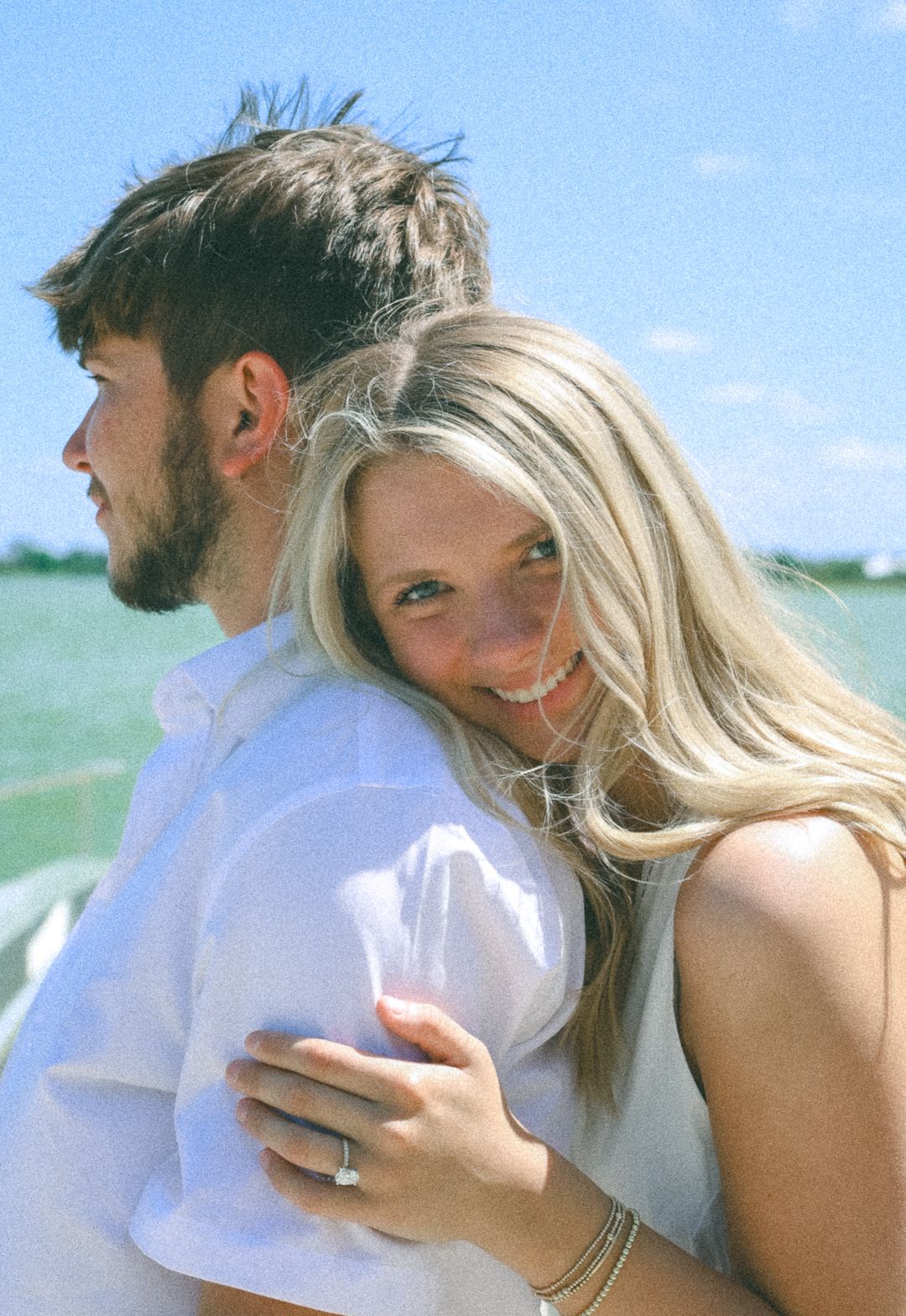 A smiling blonde woman hugs a dark-haired bearded man in white, outdoors near water on a sunny day.