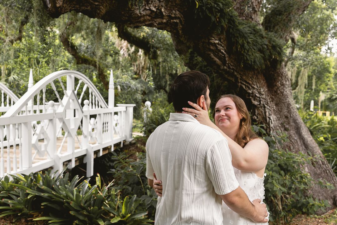 Couple embracing and smiling in a lush garden with a white bridge and large oak tree backdrop.