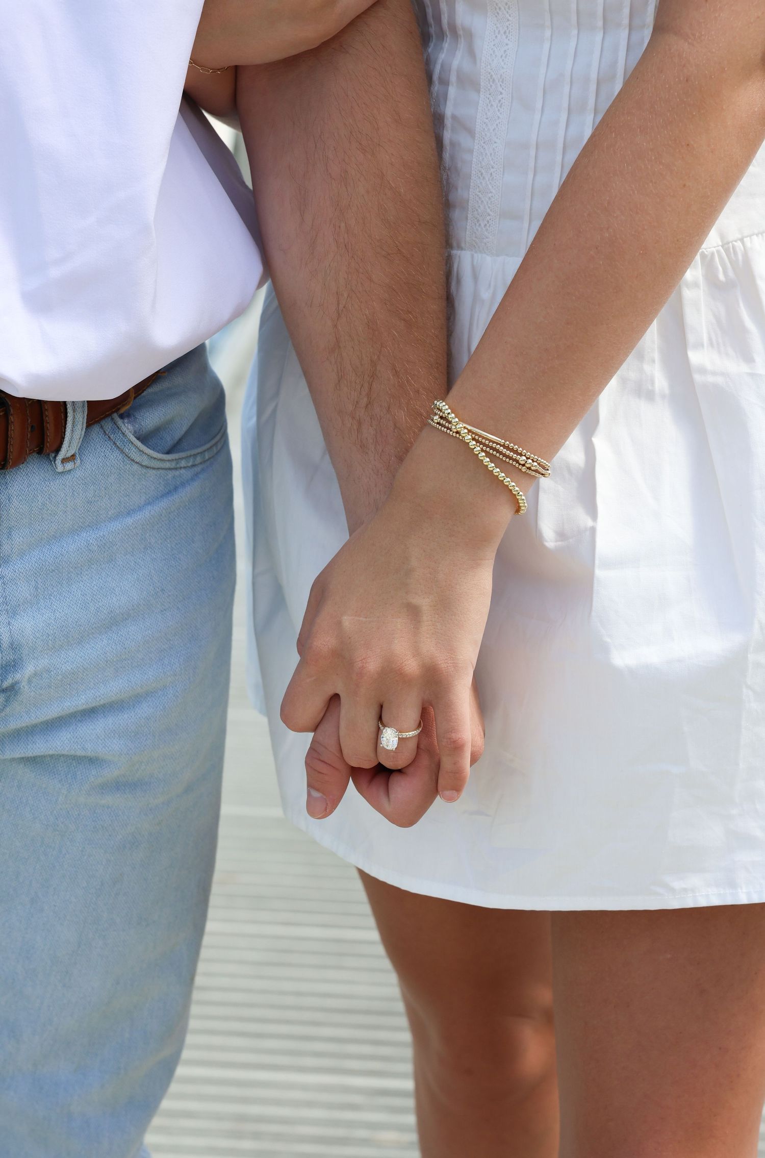 Close-up of an engaged couple holding hands, showcasing a pearl engagement ring and gold bracelets.