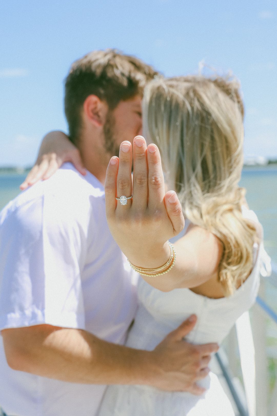 Engaged couple kissing on waterfront dock while woman displays her diamond engagement ring and gold bracelets.