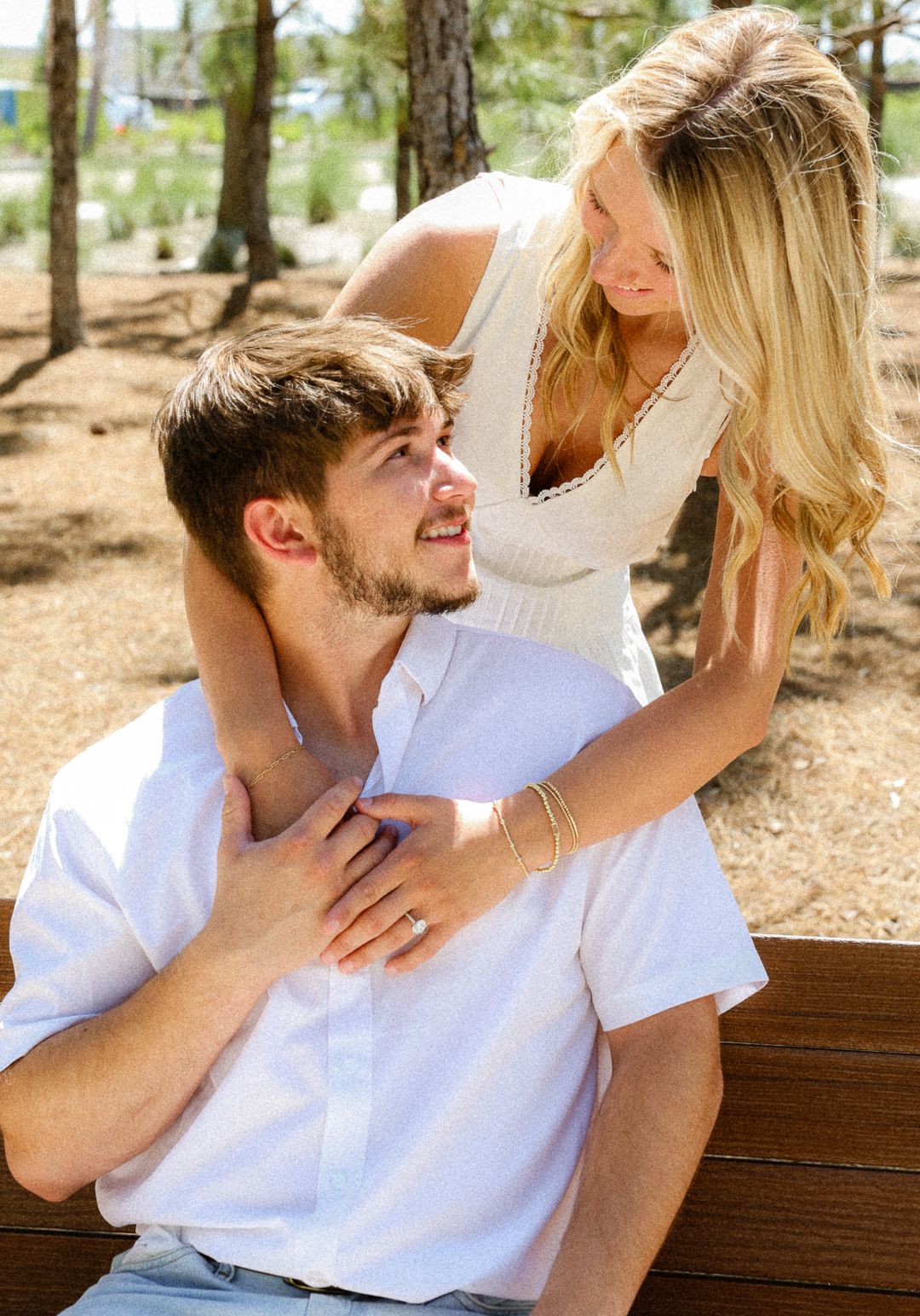 A smiling couple sharing a tender moment on a bench surrounded by pine trees outdoors.