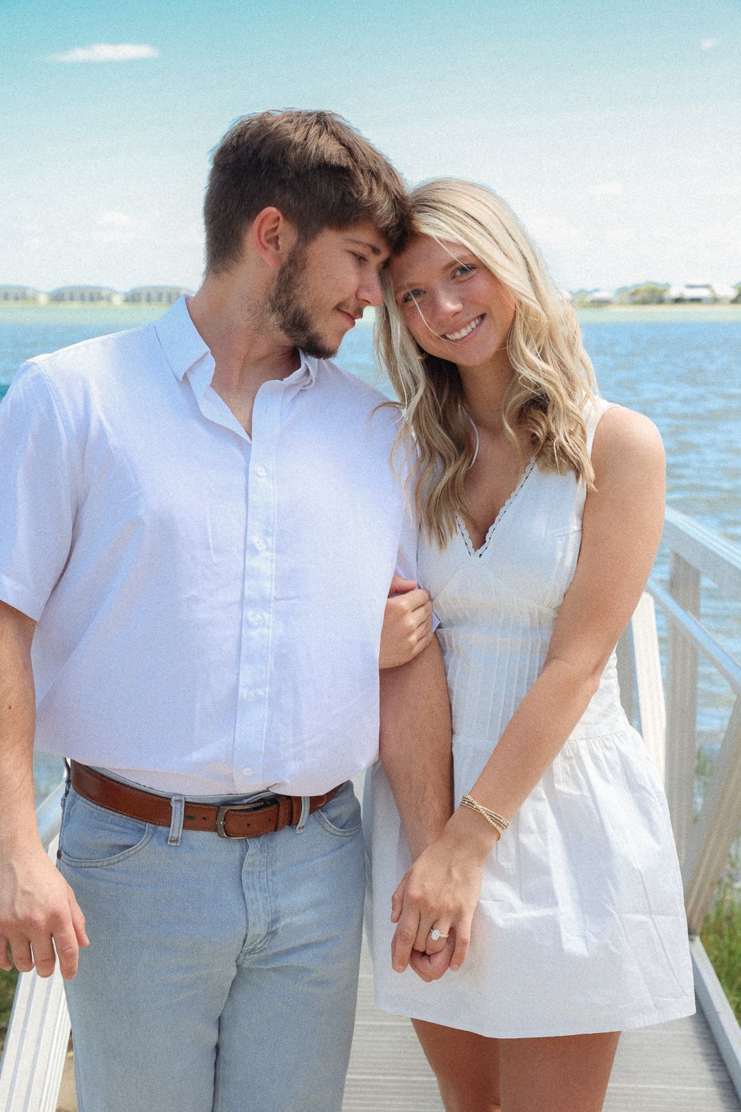 Happy engaged couple in matching white outfits embrace on sunny waterfront dock, woman smiling brightly.