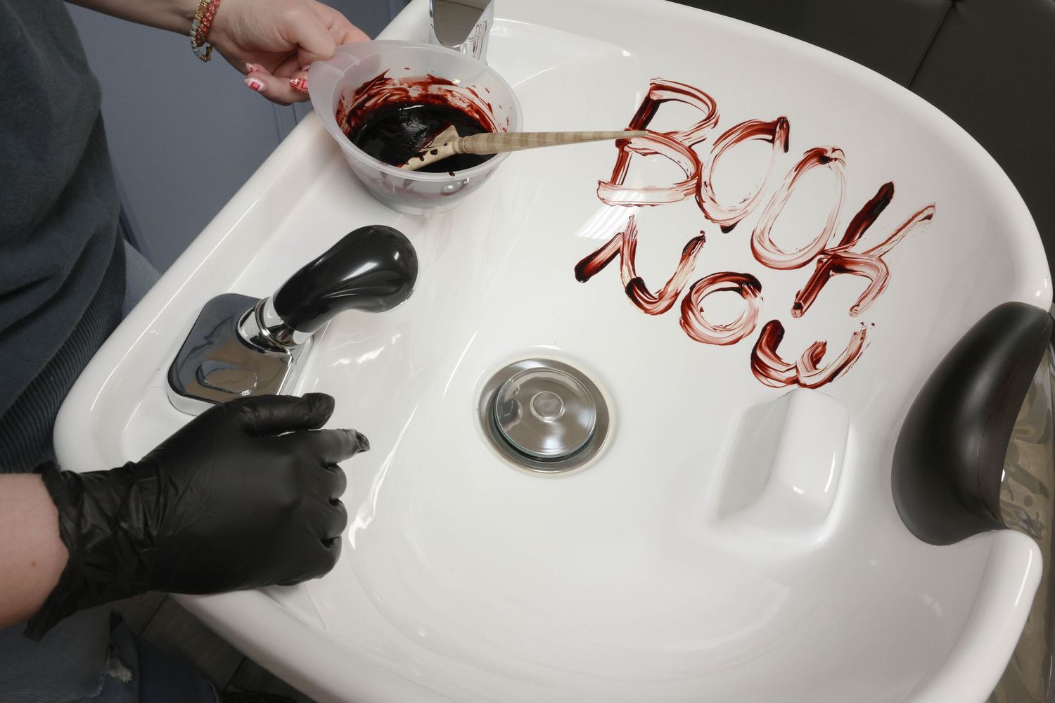 Gloved hands writing Book Now in red gel nail polish inside a white salon wash basin.