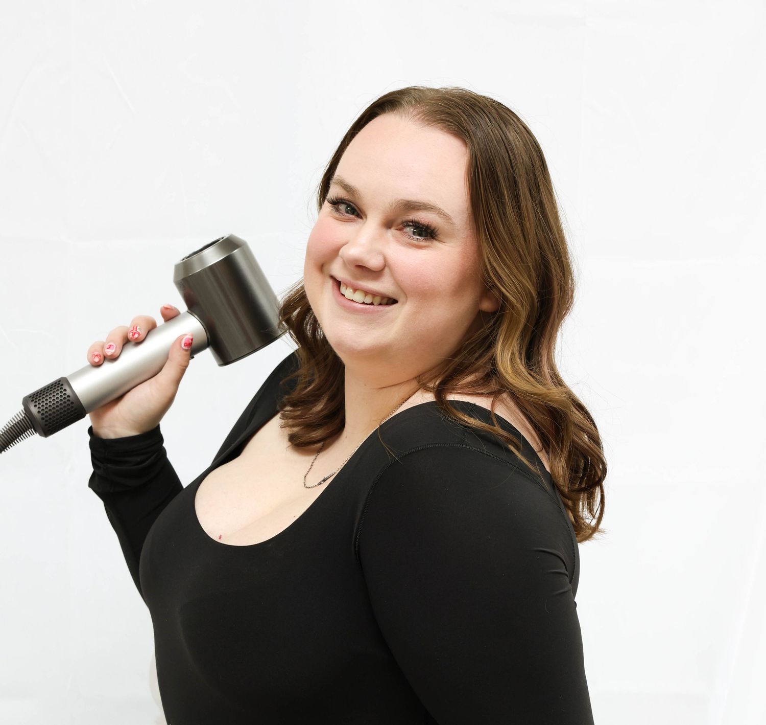 Cheerful woman in black top posing with a Dyson hair dryer against a clean white background.