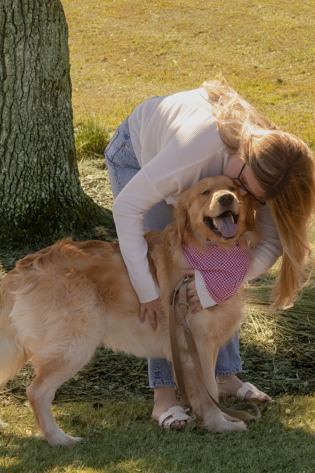 Woman with glasses affectionately hugging a happy golden retriever with pink bandana near a tree.
