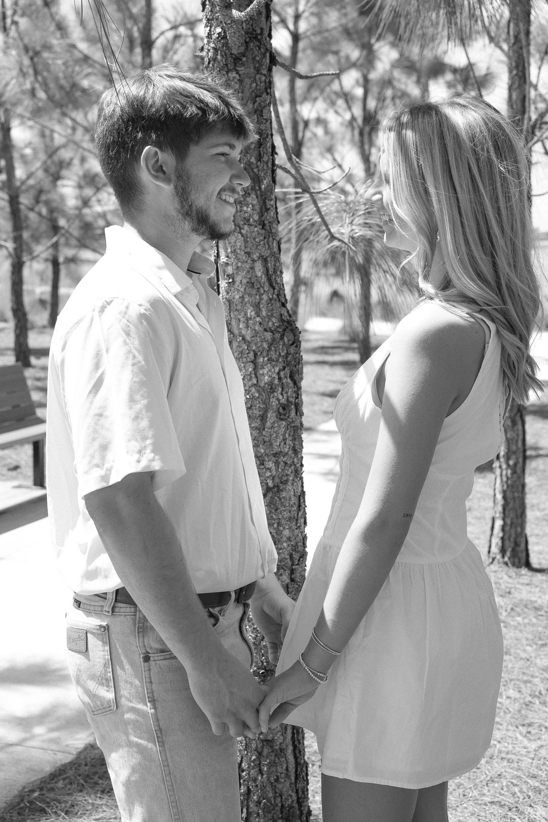 Black and white photo of smiling couple holding hands near pine tree outdoors.