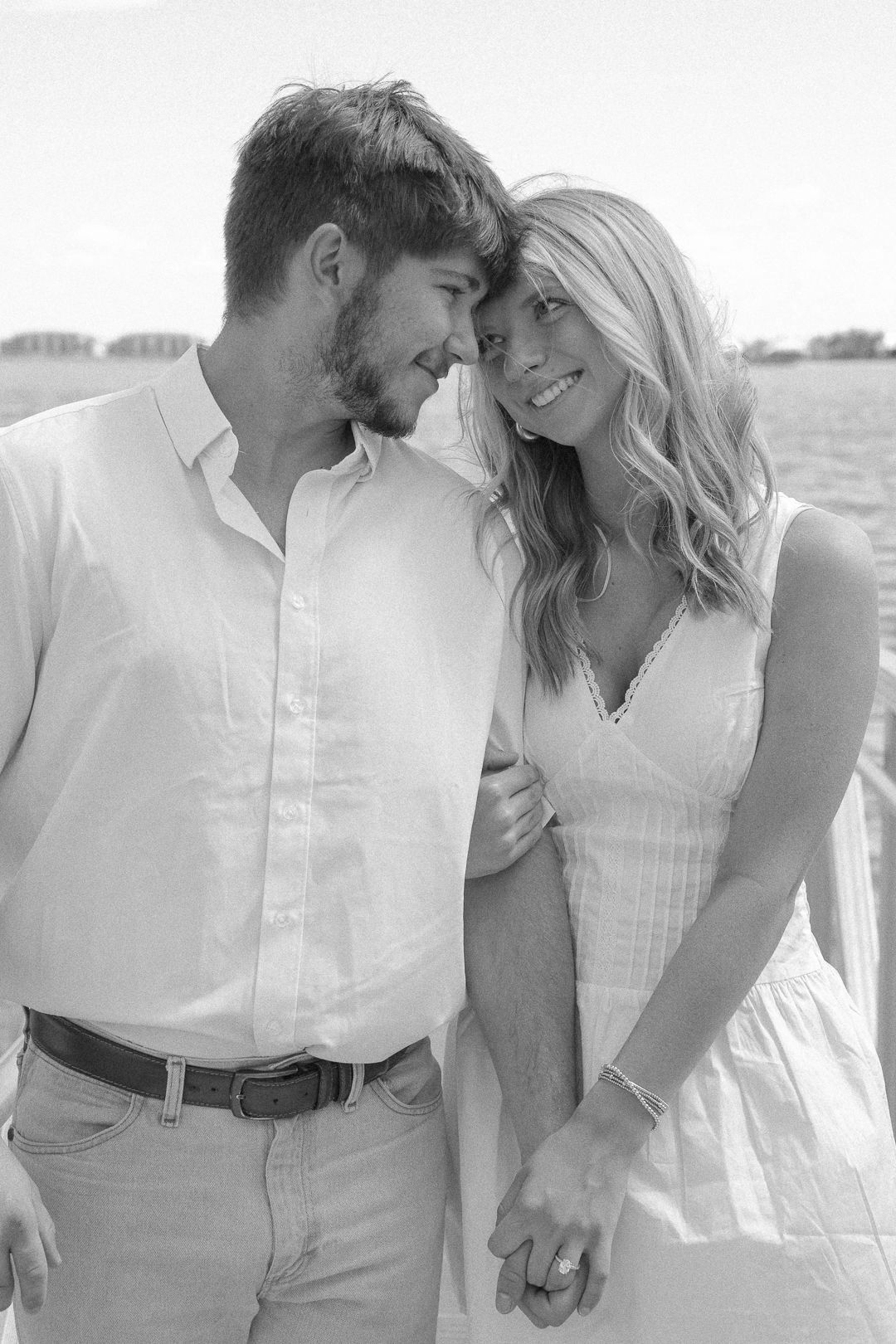 Black and white photo of engaged couple touching foreheads and smiling together near waterfront.