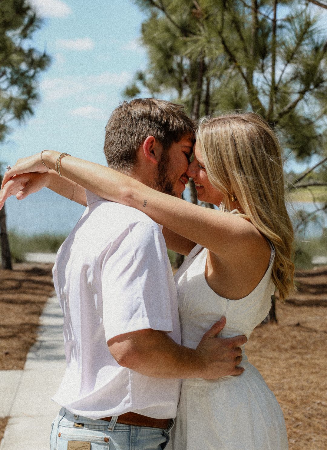 Couple embracing and touching foreheads on sunny outdoor path near pine trees.