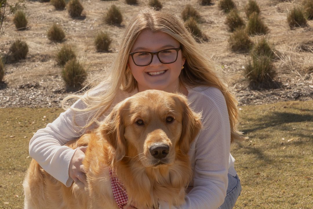 Smiling blonde girl with glasses hugging a golden retriever outdoors in a sunny field.