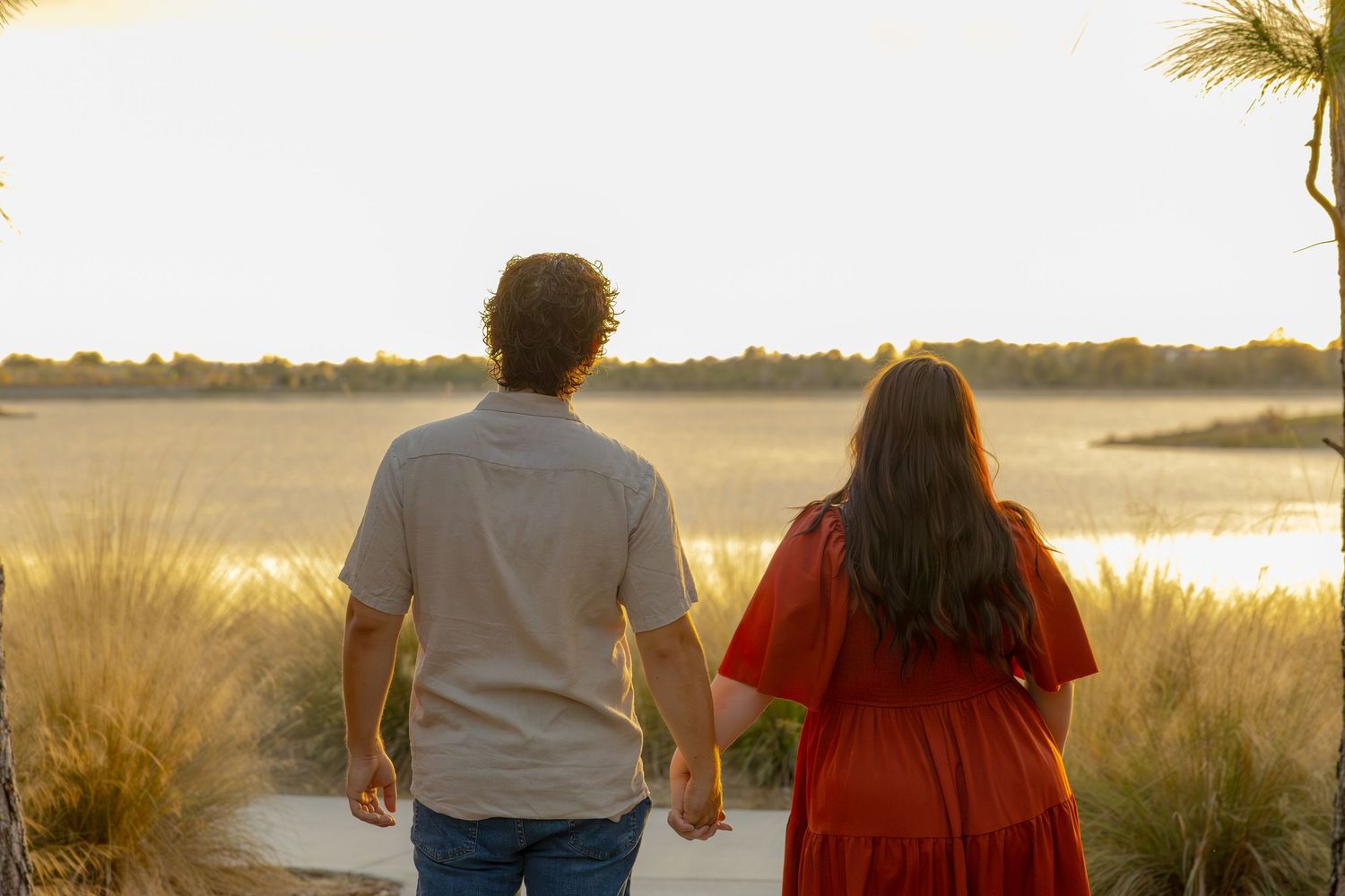 Couple holds hands walking toward a golden sunset over a marsh waterway.