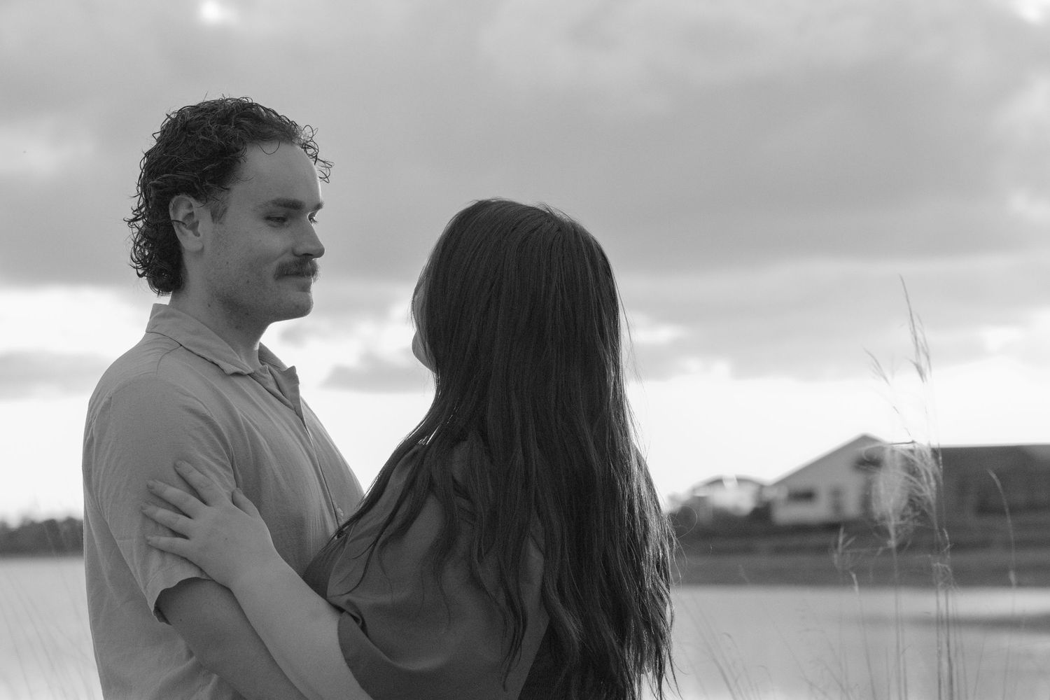 Black and white photo of a couple embracing tenderly by a serene lakeside with cloudy skies.