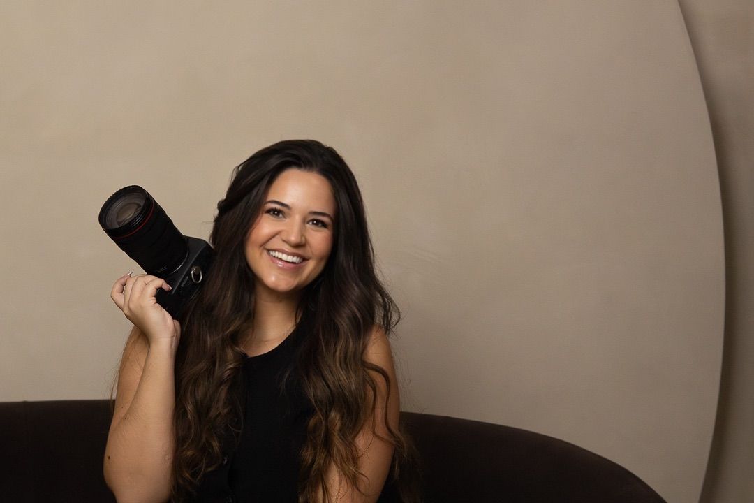 Smiling brunette woman holding a professional camera on her shoulder against a neutral beige background.