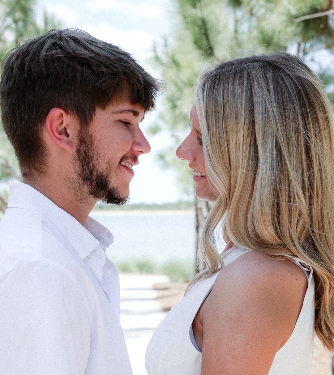 Smiling couple gazing at each other near pine trees with lake in background.