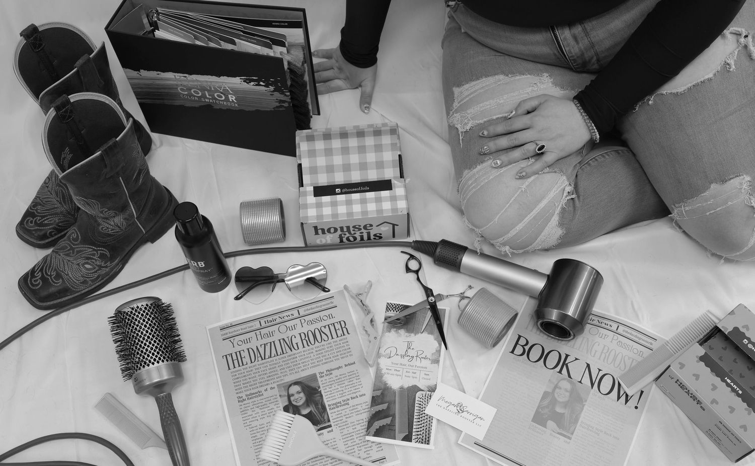 Black and white flat lay of hair styling tools, cowboy boots, magazines, and beauty products on a surface.