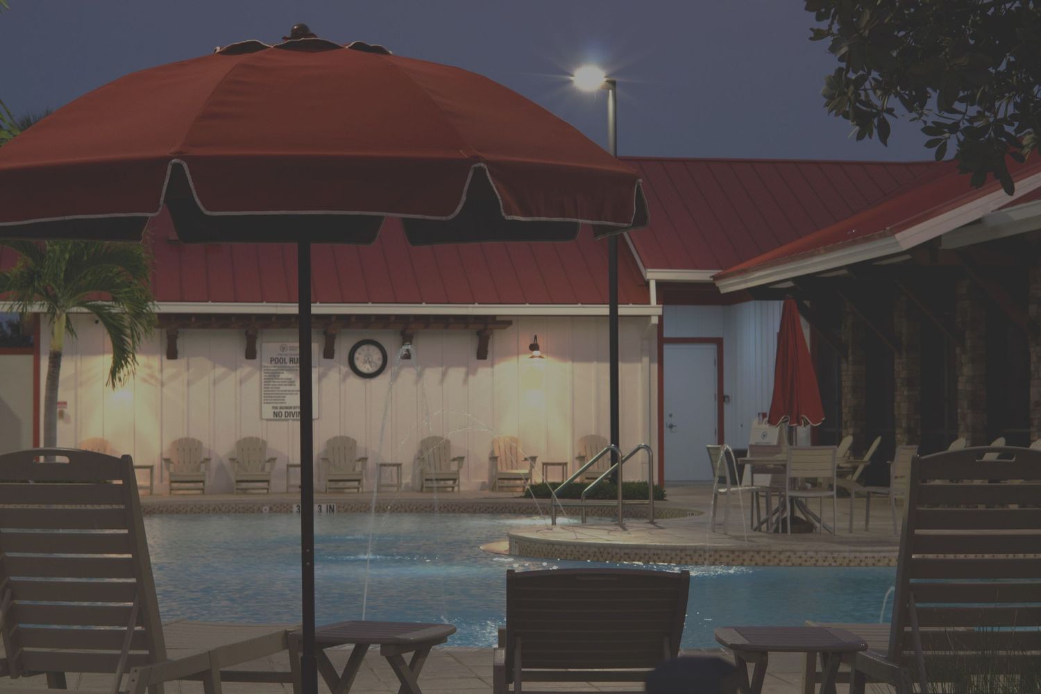 Outdoor hotel pool area at night with red umbrella, lounge chairs, and illuminated building in tropical setting.