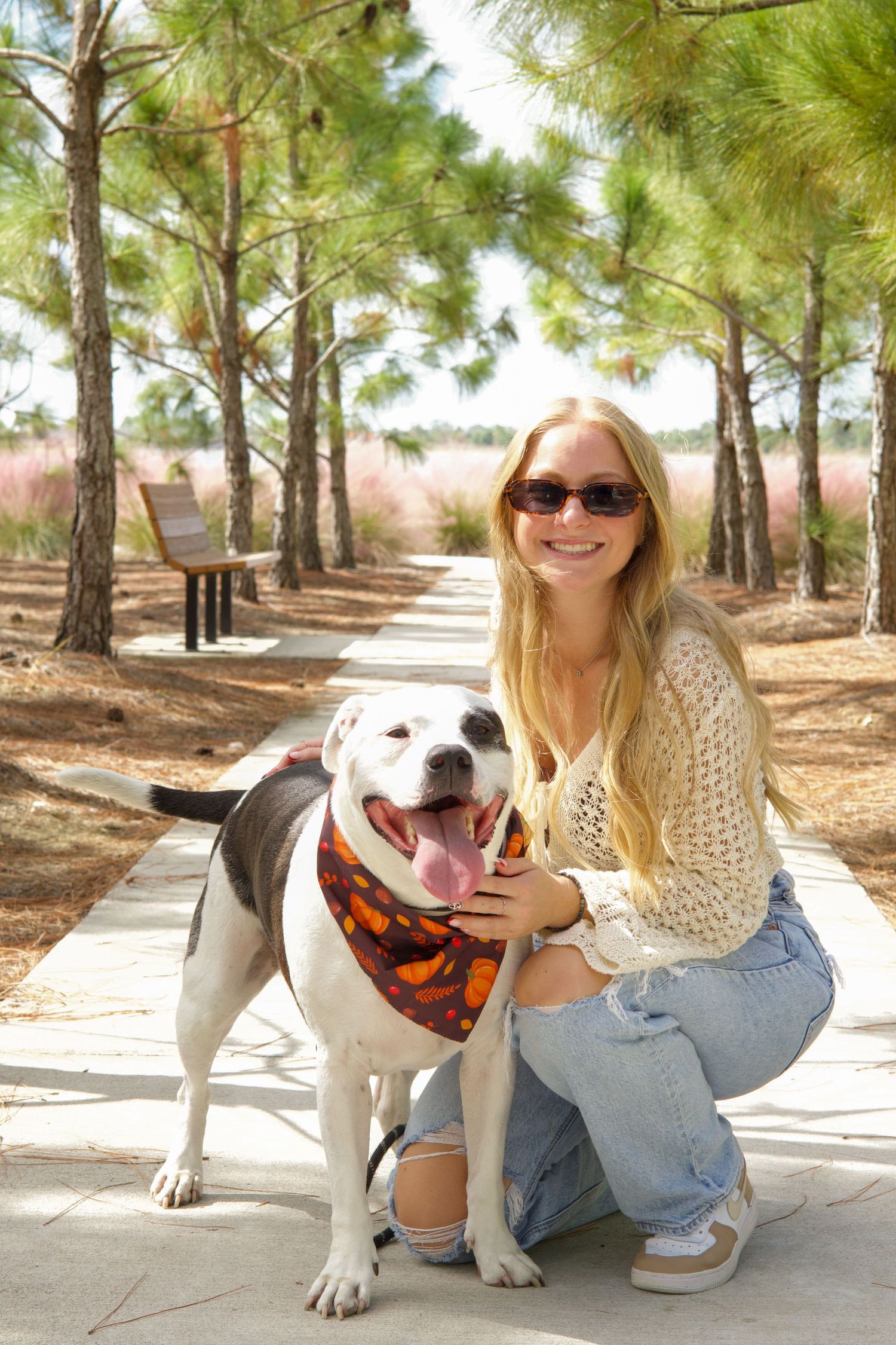 Blonde woman in sunglasses crouching with white pitbull mix dog on park path.